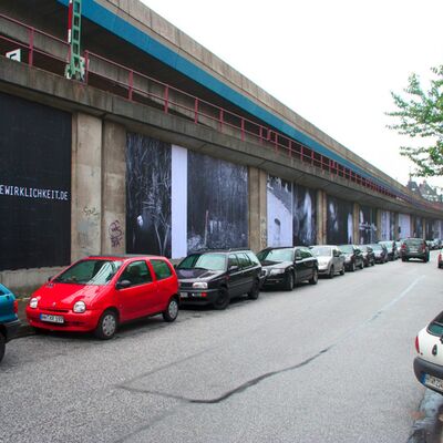 Das Bild zeigt eine Reihe von Plakaten, die entlang einer Mauer an der Straße angebracht sind.