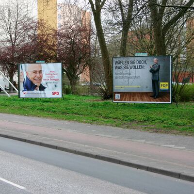 Das Bild zeigt Hauptstrasse. Im Vordergrund ist ein großes Wahlplakat zu sehen.