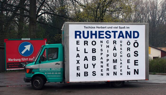 Ein kleiner Lieferwagen mit einem großen Plakat, auf dem das Wort 'Ruhestand' sowie verschiedene Synonyme abgebildet sind.