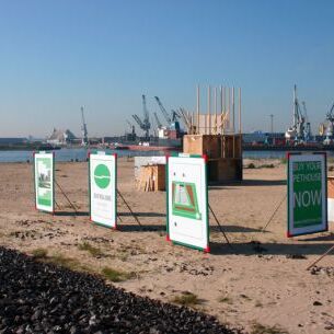 Das Bild zeigt die Elbe mit einem Sandstrand im Vordergrund, auf dem mehrere Plakate aufgestellt sind, die Informationen oder Werbung präsentieren.